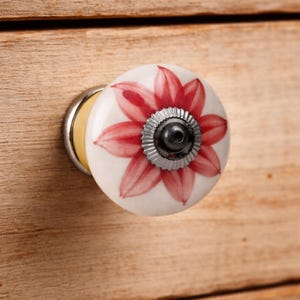 May include: A round ceramic cabinet knob featuring a white base with a painted red flower design. The center of the floral pattern is accented by a small, dark grey, ridged metal circular plate with a central screw hole for mounting purposes.