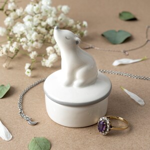 May include: White ceramic trinket box shaped like a polar bear with a gray lid. The box is sitting on a beige surface with a silver chain necklace and a ring with a purple gemstone.