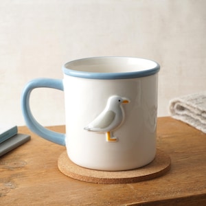 Pode incluir: Caneca de cerâmica branca com alça e borda azuis, com um design de gaivota em 3D. A caneca está sobre um descanso de copo redondo de cortiça. A alça e a borda azuis adicionam um toque de cor.