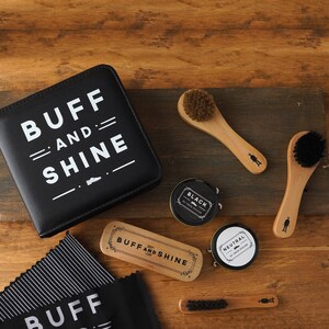 Pode incluir: Um kit de cuidados para sapatos de couro preto com as palavras "Buff and Shine" em letras brancas. O kit inclui duas latas de graxa para sapatos, uma preta e uma neutra, duas escovas de madeira, um calçador de madeira e um pano preto.