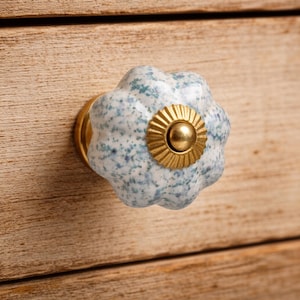 May include: A round, scalloped ceramic drawer knob featuring a white base with blue and grey speckles. The center of the knob is adorned with a circular, ridged brass-toned metal plate and a smooth, raised brass-toned central dome.