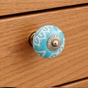 Puede incluir: Un pomo de cajón redondo de cerámica turquesa con un patrón geométrico blanco y un detalle central de metal plateado. El pomo está fijado a una superficie de madera de color marrón claro con una veta horizontal visible.