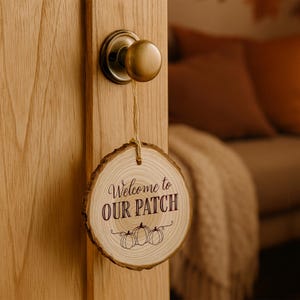 May include: A wooden door hanger with the text "Welcome to Our Patch" and three pumpkin illustrations. The round sign is made from a slice of wood and hangs from a door knob.