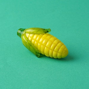 May include: A glass corn cob figurine with a yellow body and green husk. The figurine is sitting on a green surface.