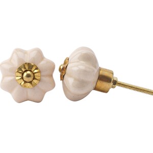 Pode incluir: Dois botões de armário de cerâmica branco-queimado com um detalhe dourado. O botão à direita tem um parafuso para fixar a uma porta de armário.