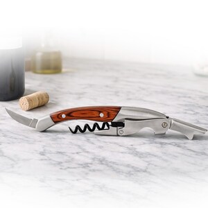 May include: A stainless steel and wood wine opener with a corkscrew, bottle opener, and small knife. The opener has a brown wood handle with silver rivets and a black corkscrew. A cork and a bottle are in the background.