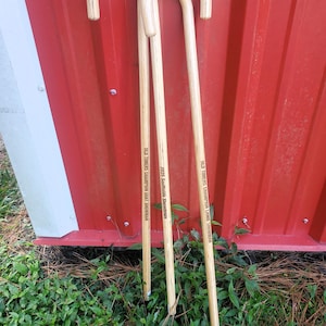 May include: Three wooden shepherd's crooks with curved handles. The crooks are light brown with text that reads "OLD TIMERS CHAMPION GOAT SHOWMAN" and "2025 Southside Showdown". The background is a red wall and green grass.