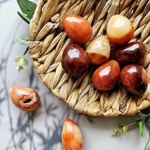 Może przedstawiać: Kolekcja wypolerowanych kamieni szlachetnych w kształcie jajka w odcieniach pomarańczu, czerwieni i brązu, ułożonych w wiklinowym koszu. Kamienie różnią się kolorem i wzorem, niektóre wykazują wewnętrzne formacje krystaliczne. Zielone liście dodają naturalnego akcentu.