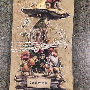 May include: A decorative journal featuring a mushroom and gnome illustration. The cover shows a large mushroom with a house, a gnome, and the word "imagine". The journal is tied with twine and has a textured, brown background.