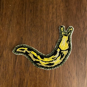 May include: A yellow and green embroidered patch of a slug with a black outline.