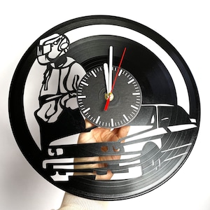May include: Black vinyl record clock featuring a silhouette of a person wearing a helmet and a car. The clock face is white with black numbers and hands.