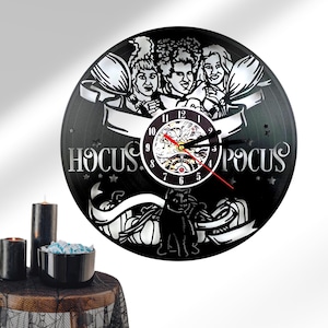 May include: Black vinyl record clock featuring the Sanderson sisters from the movie Hocus Pocus. The clock has a black background with a white clock face and silver hands. The clock is surrounded by a black frame with the words "Hocus Pocus" written in white.