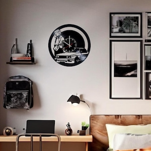 May include: A black vinyl record clock featuring a silhouette of a person driving a car. The clock is mounted on a white wall with a wooden shelf above it. There is a lamp and a desk with a laptop on it in the foreground.