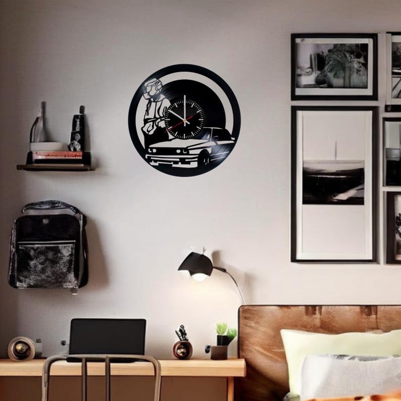 May include: A black vinyl record clock featuring a silhouette of a person driving a car. The clock is mounted on a white wall with a wooden shelf above it. There is a lamp and a desk with a laptop on it in the foreground.
