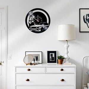 May include: A black and white vinyl record clock featuring a silhouette of a person driving a car. The clock is mounted on a white wall above a white dresser with two drawers.