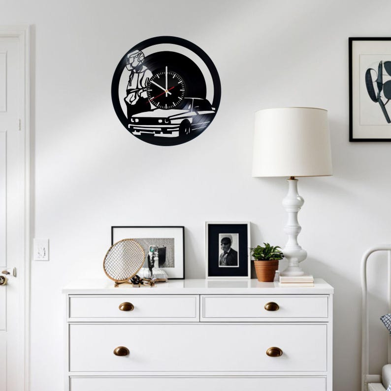May include: A black and white vinyl record clock featuring a silhouette of a person driving a car. The clock is mounted on a white wall above a white dresser with two drawers.