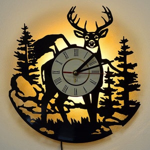 May include: A black vinyl record clock with a silhouette of a deer standing in a forest. The clock face is white with black Roman numerals and black hands. The clock is illuminated from behind, creating a warm glow.