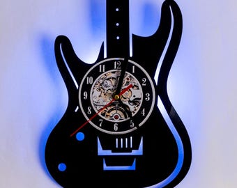 Könnte beinhalten: Schwarze, gitarrenförmige Wanduhr mit weißem Zifferblatt und silbernem Zahnrad-Design. Die Uhr hat einen roten Sekundenzeiger und blaue LED-Leuchten.