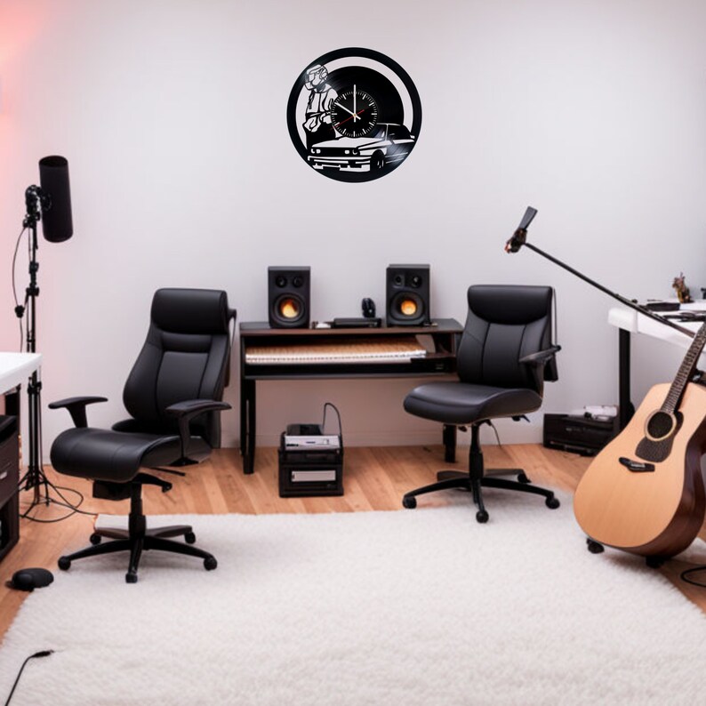 May include: A black and white wall clock with a silhouette of a person holding a guitar and a car. The clock is hanging on a white wall in a room with black office chairs, a desk, and a guitar.