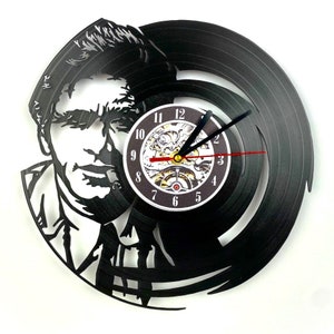 May include: A black vinyl record clock featuring a silhouette of a man's face. The clock face is a black circle with white numbers and a gold-colored clock mechanism.