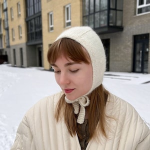 May include: A woman wearing a white knitted bonnet with a white quilted jacket. The jacket has a tie at the waist and a quilted pattern. The woman is standing in a snowy area.