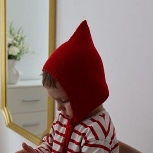May include: A bright red knitted hood with a pointed top. The hood is worn by a child wearing a red and white striped long-sleeve shirt. The child is sitting on a wooden chair and holding a small toy.