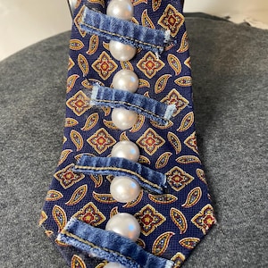 Puede incluir: Una corbata azul marino con un estampado de cachemira en rojo, dorado y blanco. La corbata está adornada con grandes cuentas blancas con forma de perla y detalles de mezclilla.