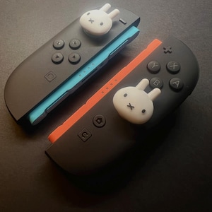 May include: Two black Nintendo Switch Joy-Con controllers with white bunny-shaped thumb grips. One controller has a blue accent, the other an orange accent. The controllers have black buttons and directional pads.