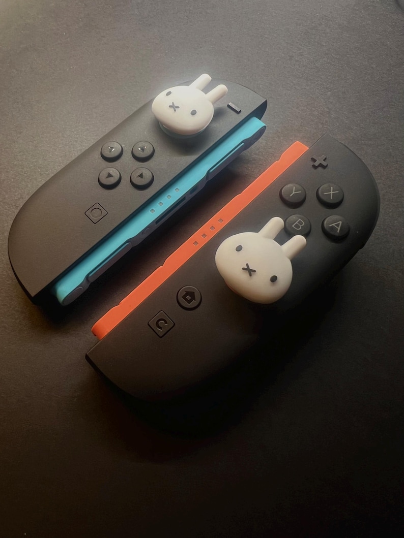 May include: Two black Nintendo Switch Joy-Con controllers with white bunny-shaped thumb grips. One controller has a blue accent, the other an orange accent. The controllers have black buttons and directional pads.