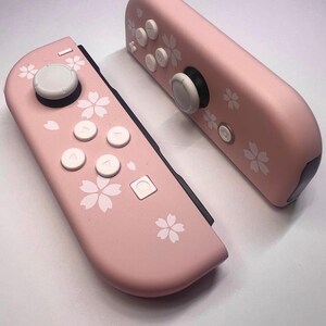 以下が含まれることがあります： 白ボタンと花の模様が入ったピンク色のゲームコントローラー2つ。ゲーム機用に設計されたコントローラーです。コントローラーは、白い花の模様と白いボタンが付いた明るいピンク色をしています。