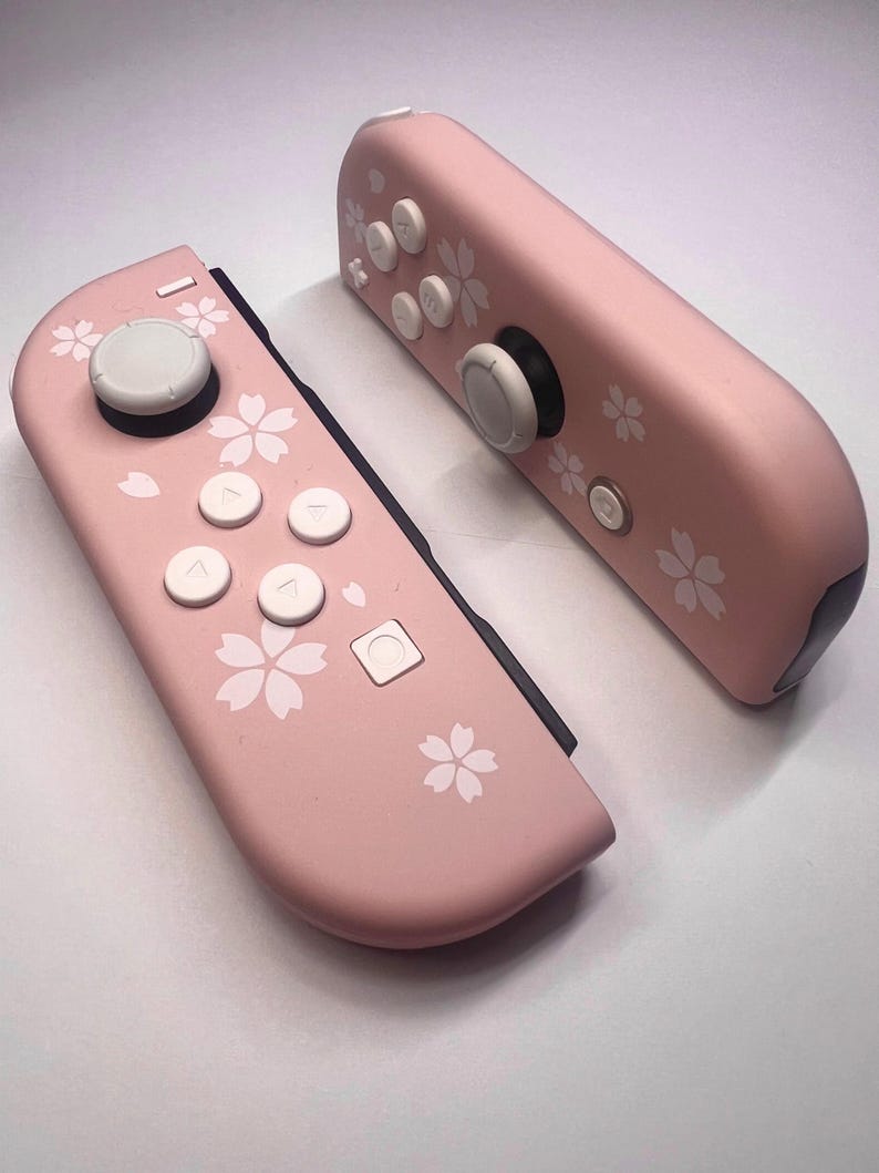 以下が含まれることがあります： 白ボタンと花の模様が入ったピンク色のゲームコントローラー2つ。ゲーム機用に設計されたコントローラーです。コントローラーは、白い花の模様と白いボタンが付いた明るいピンク色をしています。