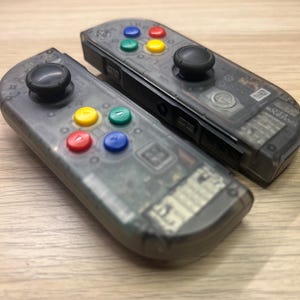 May include: Two transparent gray game controllers with black joysticks and colorful buttons. The buttons are red, yellow, green, and blue. The controllers are lying on a light brown wooden surface.