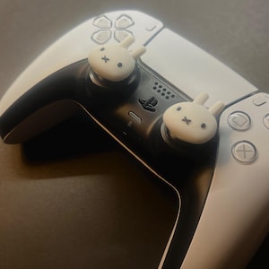 May include: A white and black PlayStation 5 controller with two white bunny-shaped thumb grips. The controller has a black centre section and white side panels with various buttons and a touchpad. The bunny grips have black facial features.