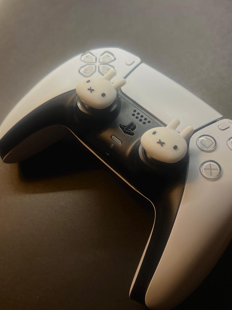 May include: A white and black PlayStation 5 controller with two white bunny-shaped thumb grips. The controller has a black centre section and white side panels with various buttons and a touchpad. The bunny grips have black facial features.