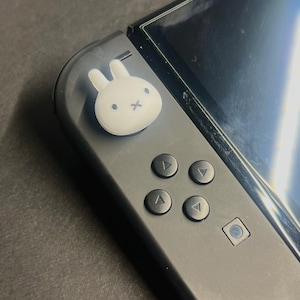 May include: A black Nintendo Switch controller with a white bunny-shaped thumb grip. The controller has four directional buttons and a small square button. The screen is visible on the right side.