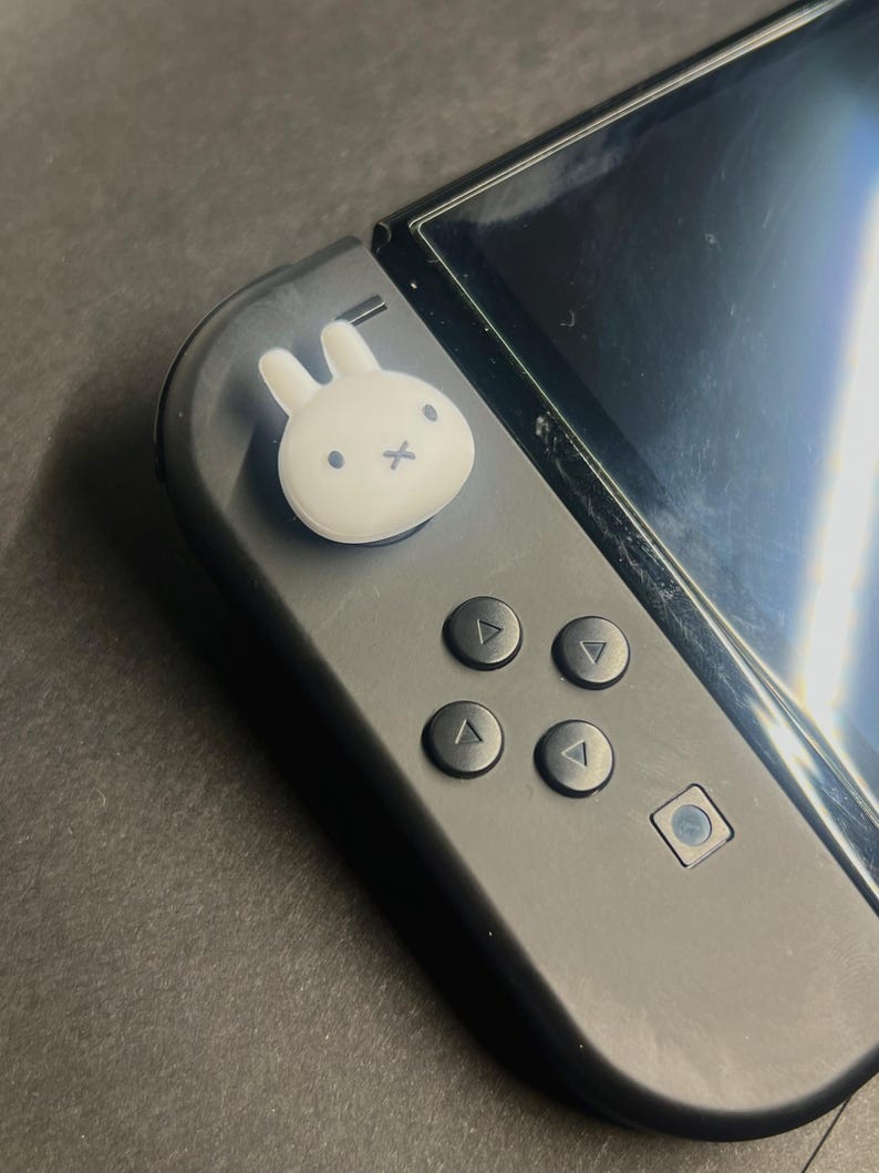 May include: A black Nintendo Switch controller with a white bunny-shaped thumb grip. The controller has four directional buttons and a small square button. The screen is visible on the right side.