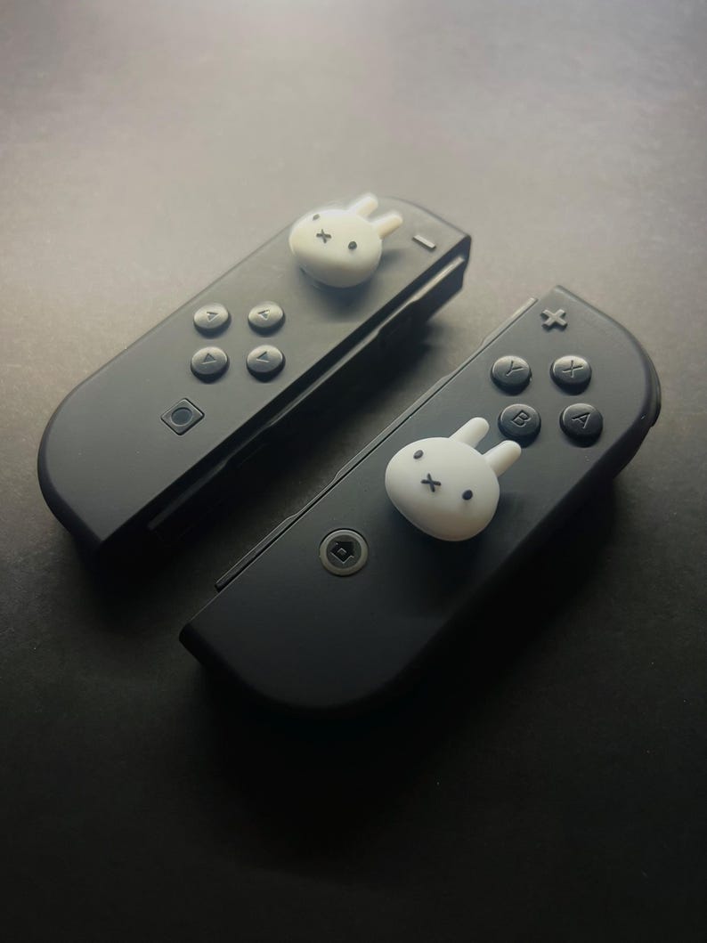 May include: Two black game controllers with white bunny-shaped decorations. The controllers have buttons with letters and symbols. The bunny decorations are on the top of each controller.
