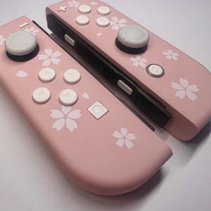 Op de afbeelding: Twee roze gamecontrollers met witte knoppen en een bloemenpatroon. De controllers hebben een lichtroze basiskleur met witte kersenbloesempatronen. De knoppen zijn wit en de joysticks zijn grijs.