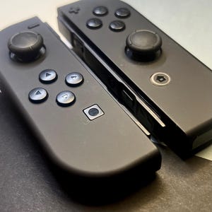 May include: Two black game controllers with multiple buttons and joysticks. The controllers are angled towards each other, with a focus on the buttons and the overall design. The controllers are likely for a gaming console.