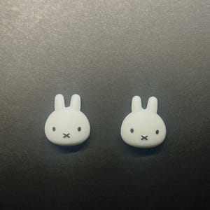 May include: Two white bunny-shaped decorative objects with black facial features. The bunnies have a minimalist design with rounded bodies and upright ears. The objects are set against a dark background.