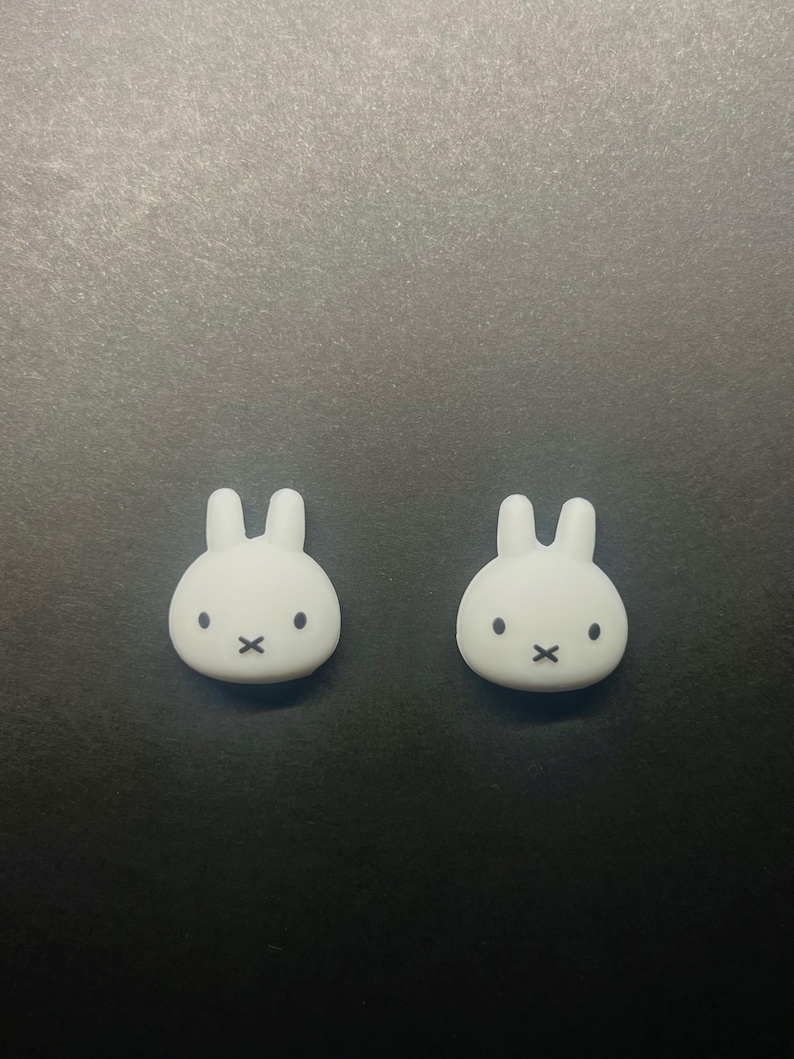 May include: Two white bunny-shaped decorative objects with black facial features. The bunnies have a minimalist design with rounded bodies and upright ears. The objects are set against a dark background.