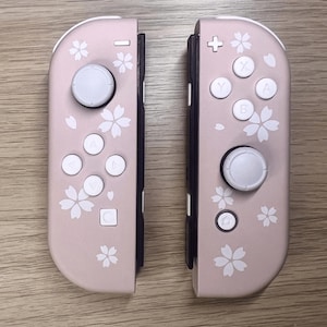 以下が含まれることがあります： ピンク色のNintendo Switch Joy-Conコントローラー2つ。各コントローラーには白いボタンと白いジョイスティックが付いています。コントローラーは白い桜のデザインで装飾されています。