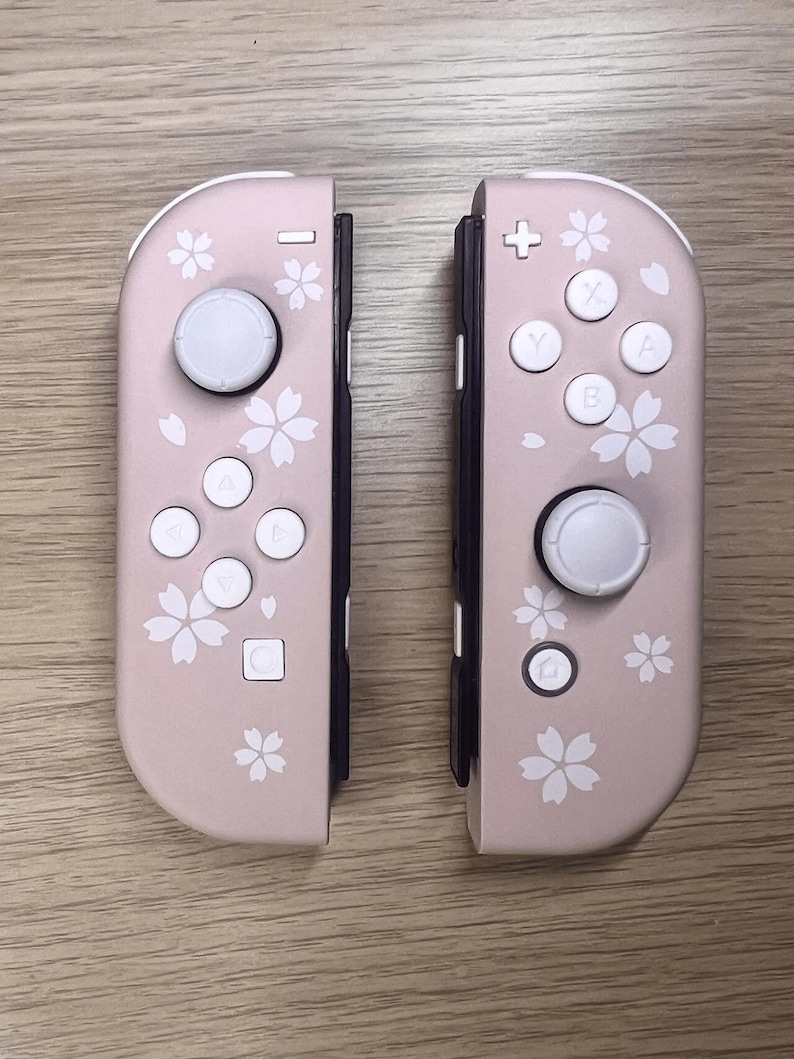 以下が含まれることがあります： ピンク色のNintendo Switch Joy-Conコントローラー2つ。各コントローラーには白いボタンと白いジョイスティックが付いています。コントローラーは白い桜のデザインで装飾されています。