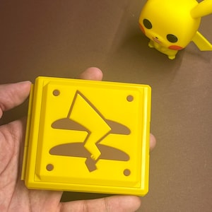 Könnte beinhalten: Ein gelber, quadratischer Behälter mit einem Blitzdesign, der in einer Hand gehalten wird. Eine gelbe Pikachu-Figur befindet sich im Hintergrund. Der Behälter hat einen Klappdeckel und vier kleine, dunkelbraune Kreise an den Ecken.