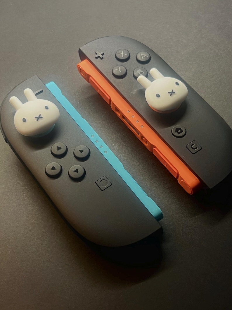 May include: Two black Nintendo Switch Joy-Con controllers with white bunny-shaped thumb grips. One controller has an orange rail and the other has a blue rail. The controllers have black buttons and directional pads.