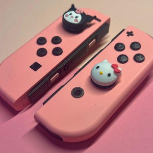 Può includere: Due controller Joy-Con rosa per Nintendo Switch con pulsanti neri e un binario nero. Un Joy-Con ha un ciondolo con personaggio nero e l'altro un ciondolo Hello Kitty. Lo sfondo è rosa.