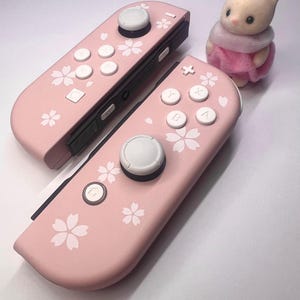 以下が含まれることがあります： 白いボタンと花の模様が付いた、2つのピンク色のゲームコントローラー。ピンクのスカーフとドレスを着た小さな白いウサギのフィギュアがコントローラーの隣にあります。コントローラーはゲーム機用です。