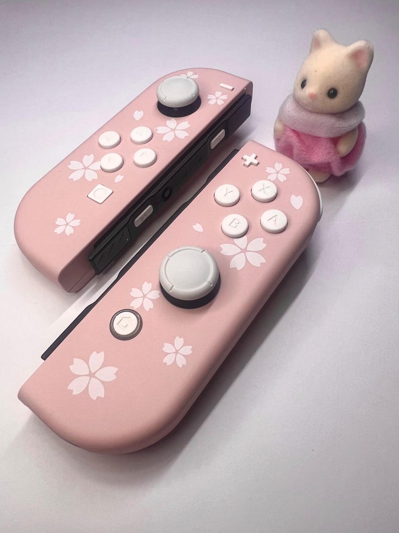 以下が含まれることがあります： 白いボタンと花の模様が付いた、2つのピンク色のゲームコントローラー。ピンクのスカーフとドレスを着た小さな白いウサギのフィギュアがコントローラーの隣にあります。コントローラーはゲーム機用です。
