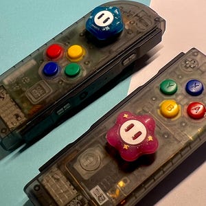 Puede incluir: Dos mandos de juego transparentes con botones coloridos. Uno tiene un botón azul con un diseño de cara blanca, y el otro tiene un botón rosa con un diseño de cara blanca y estrellas doradas.