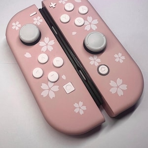 以下が含まれることがあります： ピンク色のNintendo Switch Joy-Conコントローラー。白いボタンと花の模様が特徴です。 コントローラーは明るいピンク色で、白い桜の花のパターンが施されています。 ボタンは白で、ジョイスティックはグレーです。